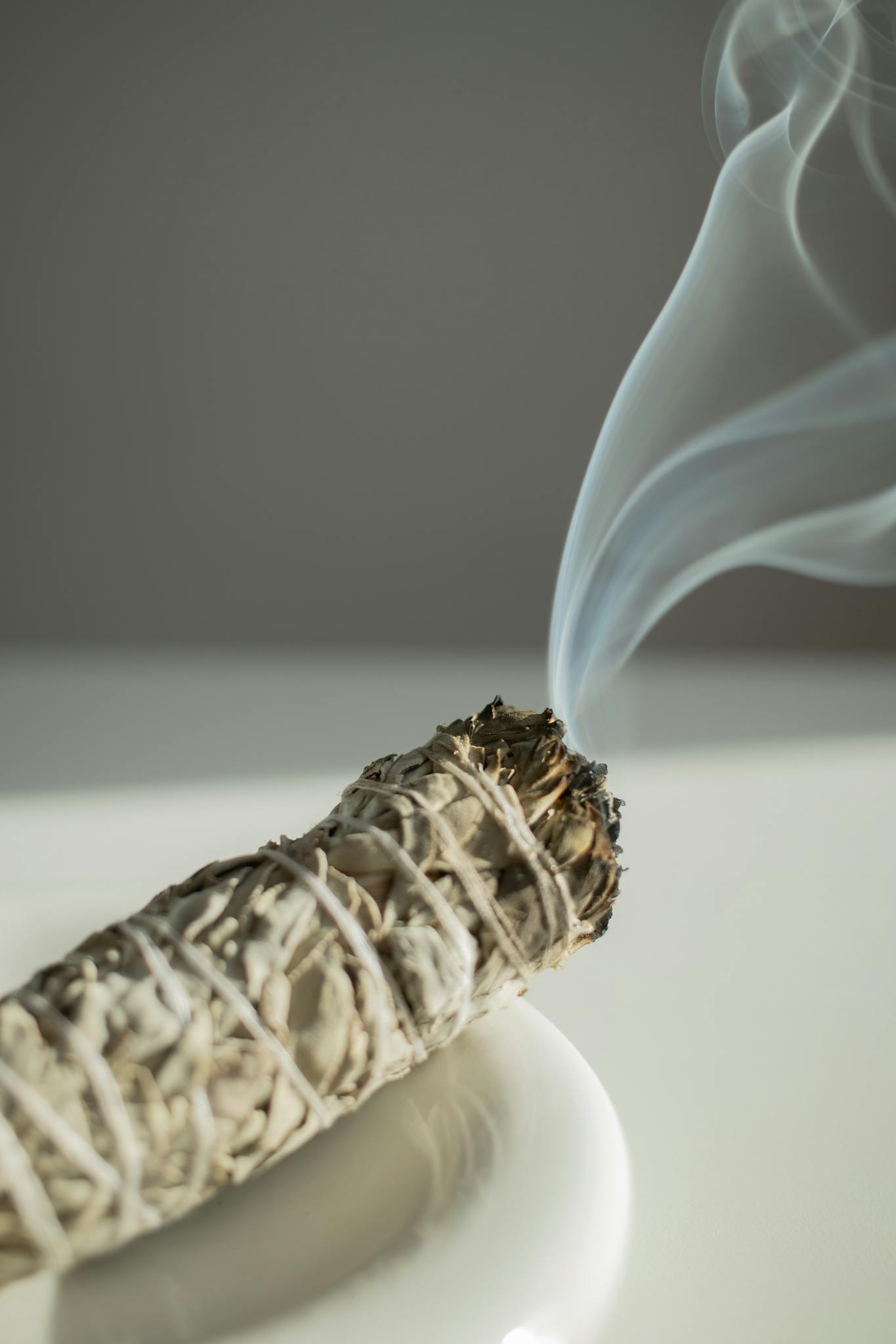A close-up of a burning sage stick emitting smoke on a white plate.