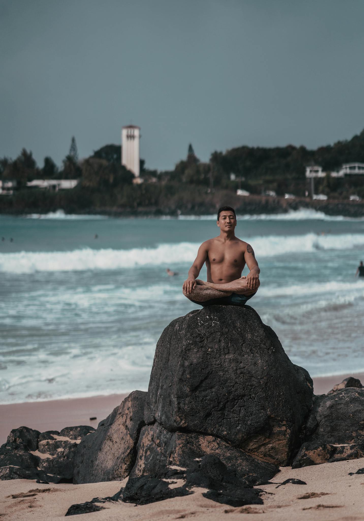 Full body of young shirtless Asian male traveler sitting in Lotus pose on rocky formation while meditating on sandy beach near waving ocean